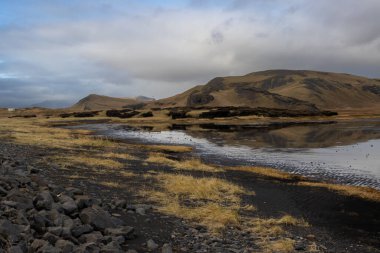 Göldeki su sonbaharda bulutlu gökyüzünü yansıtıyor. Dağlarla çevrili bir göl. Dyrholaey, Güney İzlanda.