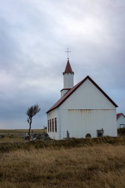 Kırmızı çatılı küçük beyaz kilise, geleneksel İzlanda kombinasyonu. Yanındaki küçük ağaç. Yoğun bulutlu gökyüzü. Çimlerin sonbahar rengi. Güney İzlanda.