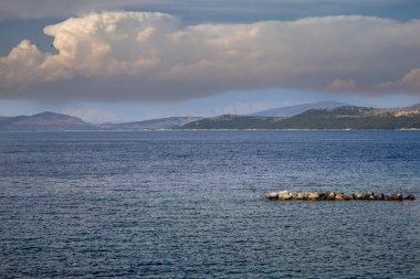 Dağlarla çevrili İyon (Akdeniz) denizinin sakin mavi suları yeşilliklerle kaplıdır. Beyaz bulutlu mavi gökyüzü dramatik bir sahne yaratıyor. Korfu, Yunanistan.