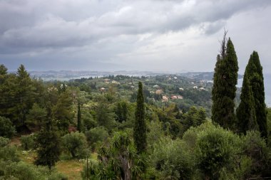 Uçurumdaki bir terastan, yeşilliklerle, ağaçlarla ve çalılarla kaplı. İyon denizi ve yoğun bulutlu yağmurlu bir gökyüzü. Yer, İmparatoriçe Sisi için bir sağlık merkezi olan Achilleon 'du. Achilleio, Korfu, Yunanistan.