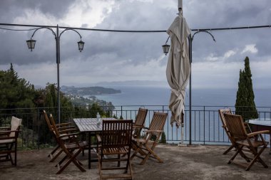 Uçurumun üzerindeki bir terastan manzara, çeşitli yeşillikler, ağaçlar ve çalılarla kaplı. İyon denizi ve yoğun bulutlu yağmurlu bir gökyüzü. Yer, İmparatoriçe Sisi için bir sağlık merkeziydi. Achilleio, Korfu, Yunanistan.