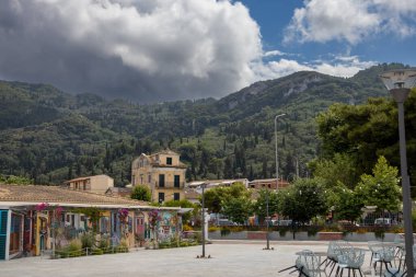 Restoranları ve barları olan deniz kenarında geniş bir gezinti alanı. Dağın altındaki eski binalar yeşillikle kaplı. Yağmurlu bulutlu mavi gökyüzü. Benitses, Korfu, Yunanistan.