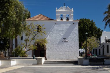 Yardım Meydanı 'ndaki (Plaza de los Remedios) park çok iyi korunmuş bir tarihi kilisenin önünde bir çan kulesiyle. Parlak mavi gökyüzü. Yaiza, Lanzarote, Kanarya Adaları, İspanya.