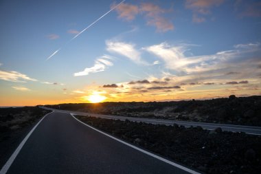 Empty good quality asphalt road on the coastline, crossing lava field. Sun on the horizon during sunset. Blue sky with colorful clouds. Salinas de Janubio, Lanzarote, Canary Islands, Spain.