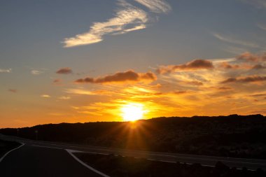 Empty good quality asphalt road on the coastline, crossing lava field. Sun on the horizon during sunset. Blue sky with colorful clouds. Salinas de Janubio, Lanzarote, Canary Islands, Spain.