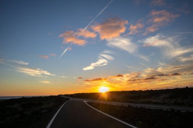 Empty good quality asphalt road on the coastline, crossing lava field. Sun on the horizon during sunset. Blue sky with colorful clouds. Salinas de Janubio, Lanzarote, Canary Islands, Spain.