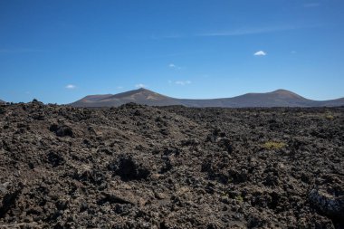 Detail of a textured dark brown to black volcanic soil. Mountain in the background. Blue sky with some white clouds. Central Lanzarote, Canary Islands, Spain.