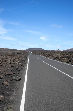 Very good quality of an empty road, surrounded by textured dark brown to black volcanic soil. Mountain in the background. Blue sky with some white clouds. Central Lanzarote, Canary Islands, Spain.