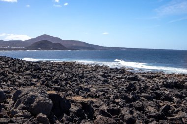 Detail of a textured dark brown to black volcanic stones on the coast of Atlantic ocean. Mountain in the background. Blue sky with some white clouds. Central Lanzarote, Canary Islands, Spain.