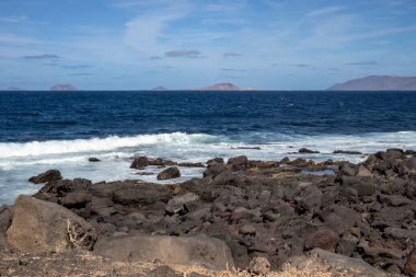 Detail of a textured dark brown to black volcanic stones on the coast of Atlantic ocean. Mountain in the background. Blue sky with some white clouds. Central Lanzarote, Canary Islands, Spain.