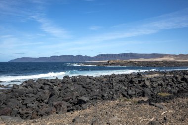 Detail of a textured dark brown to black volcanic stones on the coast of Atlantic ocean. Mountain in the background. Blue sky with some white clouds. Central Lanzarote, Canary Islands, Spain.