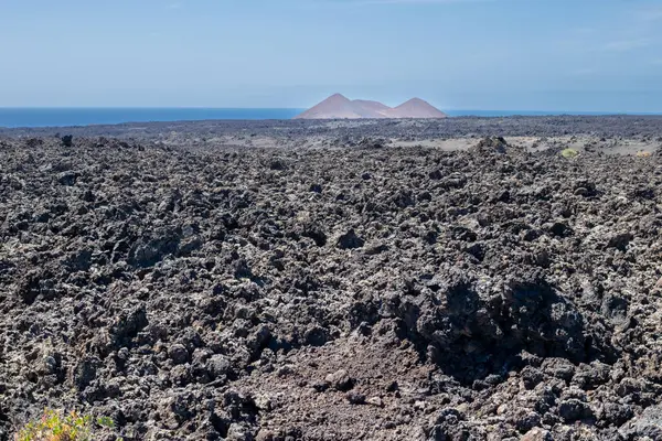 Detail of a textured dark brown to black volcanic soil. Mountain in the background. Line of Atlantic ocean. Blue sky with some white clouds. Central Lanzarote, Canary Islands, Spain.