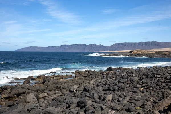 Detail of a textured dark brown to black volcanic stones on the coast of Atlantic ocean. Mountain in the background. Blue sky with some white clouds. Central Lanzarote, Canary Islands, Spain.