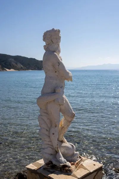 Calm and clean Ionian sea. Statue of Nereus in the harbor. Glittering water. Blue sky with light clouds. Mountains of Corfu in the background. Island Erikousa, Greece.