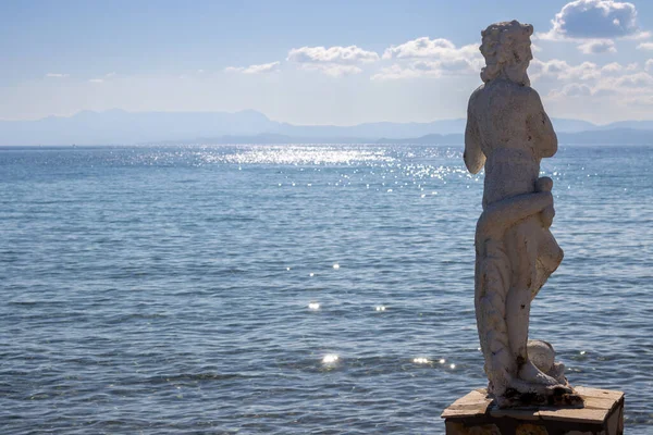 Calm and clean Ionian sea. Statue of Nereus in the harbor. Glittering water. Blue sky with light clouds. Mountains of Corfu in the background. Island Erikousa, Greece.