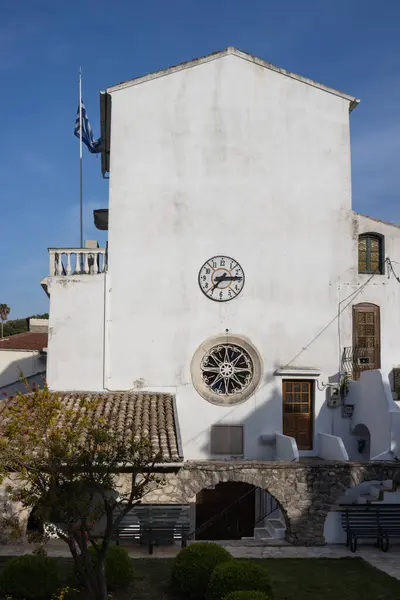 Beyaz yıpranmış cepheli ve duvar saati olan eski bir binanın yan tarafında. Güneşli bir bahar günü. Parlak mavi gökyüzü. Ön planda bahçe var. Kassiopi, Korfu, Yunanistan