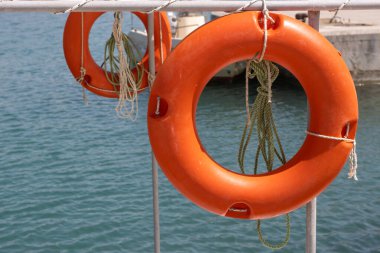 Aydınlatılmış turuncu can simidi, beyaz çitte asılı. Arka planda bir tane daha. İyon (Akdeniz) denizinin sakin suları. Roda, Korfu, Yunanistan.