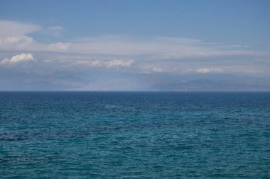 İyon (Akdeniz) denizinin sakin suları. Mavi 'den turkuaz rengine. Diğer taraftaki dağlar, kısmen bulutlarla kaplı. Sidari, Korfu, Yunanistan.