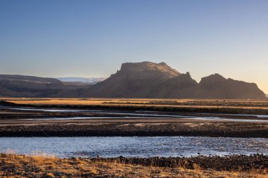 Ot ve yosun rengi sarı-yeşil bir ülke. Bir nehir çizgisi ve arka planda bir dağ. Sonbahar güneşli bir günde mavi gökyüzü. Onundarhorn, Güney İzlanda.