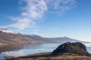 Mavi gökyüzü ve ışık bulutlarıyla güneşli bir gün. Bitki örtüsünün sonbahar renginde olduğu çayırlarla ve dağlarla çevrili bir fiyort. Eskifjordur, Doğu İzlanda.