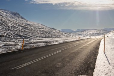 İyi bir yol, bu ada için çok tipik. Mavi gökyüzü ve sonbaharda hafif bulutlarla güneşli bir gün. Sonbaharda yol kenarında ince bir kar tabakasıyla. Fjardabyggd, Doğu İzlanda.