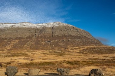 Tepesinde biraz kar olan görkemli dağ, sonbahar otlarıyla çevrili otlar. Açık beyaz bulutlu mavi gökyüzü. Neskaupstadur, Doğu İzlanda.