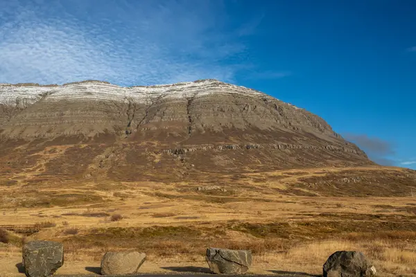 Tepesinde biraz kar olan görkemli dağ, sonbahar otlarıyla çevrili otlar. Açık beyaz bulutlu mavi gökyüzü. Neskaupstadur, Doğu İzlanda.