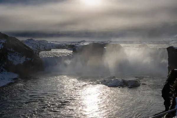 Dramatic weather at the famous waterfall. Backlight of the winter sun. The air full of steam, spray and fog. Glossy water. Godafoss, North Iceland.
