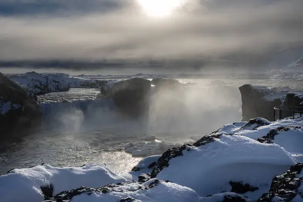 Dramatic weather at the famous waterfall. Backlight of the winter sun. The air full of steam, spray and fog. Glossy water. Godafoss, North Iceland.