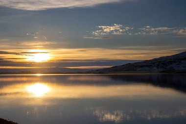 Sabahın erken saatlerinde kutup gündoğumunda. Karlı bir dağ ve bir fiyordun suyundaki yansıması. Mavi gökyüzü ve ışık bulutları. Güneş hala batıyor. Nordaustur, Kuzey-Batı İzlanda.