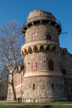 Taştan yapılmış bir şatonun sahte Gotik taklidi. Kışın sonlarında yeşil çimlerin ve birçok ağacın olduğu bir parkta. Parlak mavi gökyüzü. Kırmızı pencere panjurları. UNESCO mirası. Januv hrad, Podivin, Lednice, Morava, Çek Cumhuriyeti.