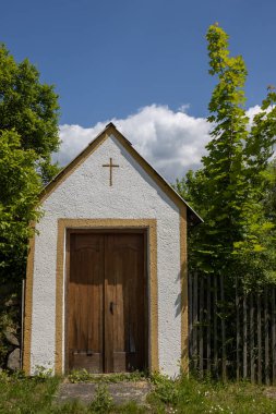 All Saints kilisesinin yanındaki bahçede haçlı küçük bir ev. Parlak yeşil ağaçlar. Beyaz bulutlu mavi gökyüzü. Dlouhomilov, Moravya, Çek Cumhuriyeti.