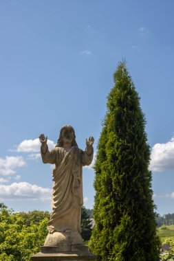 İsa 'nın heykeli mezardaki mezar taşı gibi. Tipik bir jest. All Saints kilisesinin yanındaki mezarlıkta. Yanında büyüyen yüksek parlak yeşil kozalaklı ağaç. Beyaz bulutlu mavi gökyüzü. Dlouhomilov, Moravya, Çek Cumhuriyeti.