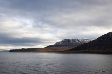 Atlantik okyanusunun sakin suları. Sahildeki yüksek dağlar arka planda. Sonbaharda yoğun bulutlu bir gökyüzü. Isafjordur, Westfjords, İzlanda Bölgesi.