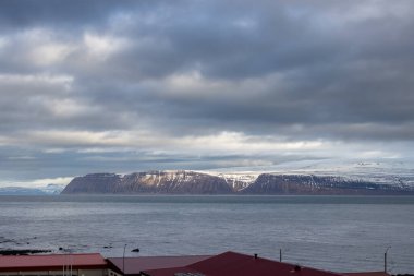 Atlantik okyanusunun sakin suları. Sahildeki yüksek dağlar arka planda. Sonbaharda yoğun bulutlu bir gökyüzü. Isafjordur, Westfjords, İzlanda Bölgesi.