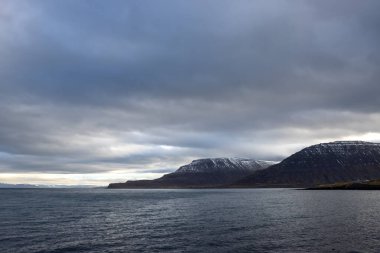 Atlantik okyanusunun sakin suları. Sahildeki yüksek dağlar arka planda. Sonbaharda yoğun bulutlu bir gökyüzü. Isafjordur, Westfjords, İzlanda Bölgesi.