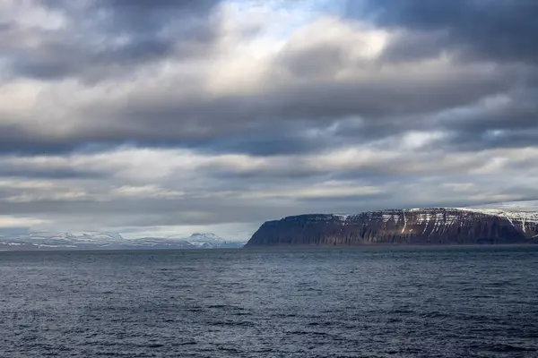 Atlantik okyanusunun sakin suları. Sahildeki yüksek dağlar arka planda. Sonbaharda yoğun bulutlu bir gökyüzü. Isafjordur, Westfjords, İzlanda Bölgesi.
