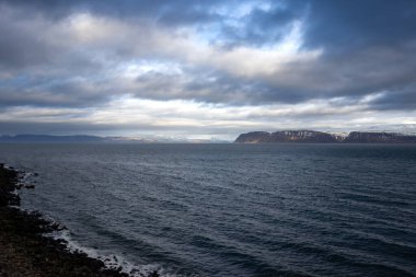 Atlantik okyanusunun sakin suları. Fiyordun diğer tarafındaki yüksek dağlarda. Ön planda Rocky Coast var. Sonbaharda yoğun bulutlu bir gökyüzü. Isafjordur, Westfjords, İzlanda Bölgesi.