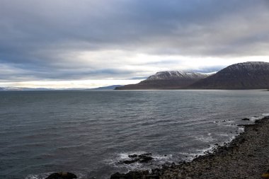 Atlantik okyanusunun sakin suları. Fiyordun diğer tarafındaki yüksek dağlarda. Ön planda Rocky Coast var. Sonbaharda yoğun bulutlu bir gökyüzü. Isafjordur, Westfjords, İzlanda Bölgesi.