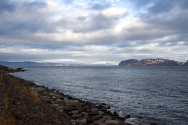 Atlantik okyanusunun sakin suları. Fiyordun diğer tarafındaki yüksek dağlarda. Ön planda Rocky Coast var. Sonbaharda yoğun bulutlu bir gökyüzü. Isafjordur, Westfjords, İzlanda Bölgesi.
