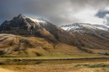 Sonbaharda kar yağışı eşliğinde görkemli bir dağ. Bulutlu bir gökyüzü, ama güneş ışınlı bir ışık. Vadideki nehir. Sydridalur, Westfjords, İzlanda Bölgesi.