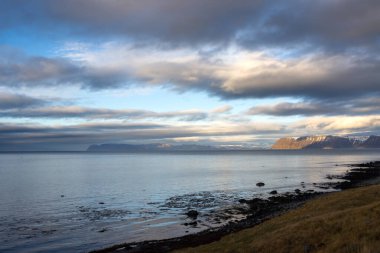 Mavi gökyüzü ve bulutlarla gün batımının başlangıcı. Fiyortta Atlantik Okyanusu 'nun Suyu. Sonbaharda biraz kar yağan görkemli tepeler. Westfjords, Isafjordur, İzlanda Bölgesi.