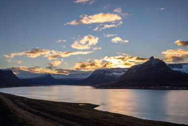 Dağlar karla kaplı. Gün batımında mavi gökyüzünde renkli bulutlar fiyorttaki Atlantik okyanusunun sakin suları. Westfjords, Isafjordur, İzlanda Bölgesi.