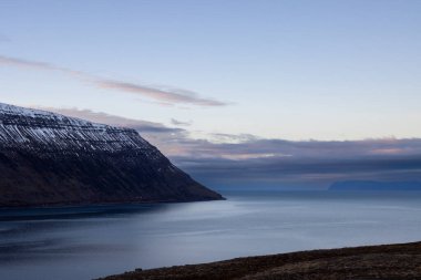 Dağlar karla kaplı. Gün batımında mavi gökyüzünde renkli bulutlar fiyorttaki Atlantik okyanusunun sakin suları. Westfjords, Isafjordur, İzlanda Bölgesi.