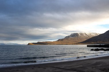 Atlantik Okyanusu 'nun karanlık volkanik kıyıları. Ufuktaki dağlar, karlı zirveler. Yoğun bulutlu gökyüzü. Isafjordur, Westfjords, İzlanda Bölgesi.