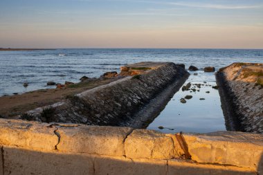 Gün doğumunda köyden denize giden kanal. Sakin bir Akdeniz. Koyu mavi su ve ufuk çizgisi. Açık bulutlu mavi-turuncu gökyüzü. Spiaggia Sibiliana, Marsala, Sicilya, İtalya.