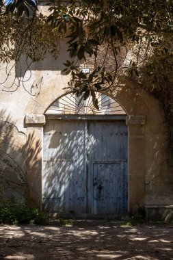 Şık, eski bir eve giriş. Girişin sağındaki eski incir ağacı güneşli bir günde gölgesini bırakıyor. Bej ön cephe ve mavi kemerli kapı. Parlak mavi gökyüzü. Ciavolo, Sicilya, İtalya.