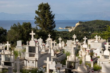 Adanın kuzeyindeki köy. Kilisenin yanındaki mezarlık, ağaçlar ve çalılarla çevrili. Arka planda İyon denizi ve Arnavutluk dağları var. Mavi bulutlu gökyüzü. Alviyotlar, Korfu, Yunanistan.