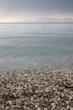 Baharın sonlarında gün batımında sakin bir deniz. Bulutlu gökyüzü. Arnavutluk kıyıları, ufukta yüksek dağlar var. Acharavi, Korfu, Yunanistan.