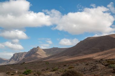 Toprağın çeşitli turuncu ve kahverengi tonlarında. Volkanik dağlar. Beyaz bulutlu mavi gökyüzü. Famara, Lanzarote, Kanarya Adaları, İspanya.
